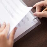 A person signs a document filled with rows of printed data while holding the pages on a wooden desk. - Ghassemi Law Group A person signs a document filled with rows of printed data while holding the pages on a wooden desk