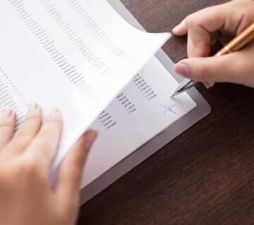 A person signs a document filled with rows of printed data while holding the pages on a wooden desk.