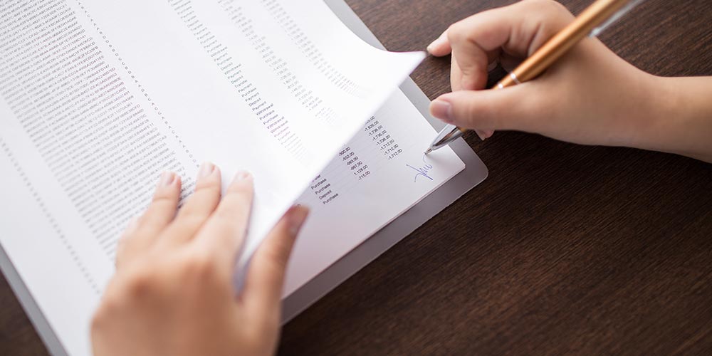 A person signs a document filled with rows of printed data while holding the pages on a wooden desk.