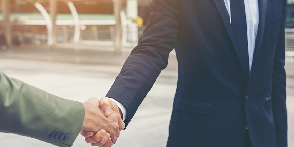 Two people in business suits shaking hands outdoors, only their arms and torsos visible.