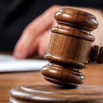 A wooden judge's gavel rests on a sound block with a person's hand and paperwork visible in the background. - Ghassemi Law Group A wooden judge's gavel rests on a sound block with a person's hand and paperwork visible in the background