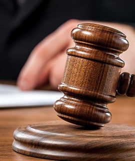 A wooden judge's gavel rests on a sound block with a person's hand and paperwork visible in the background.