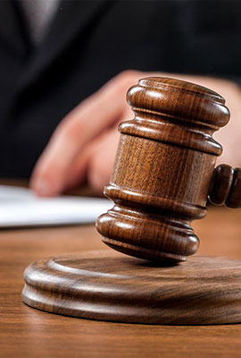 A wooden judge's gavel rests on a sound block with a person's hand and paperwork visible in the background.