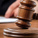 A wooden judge’s gavel rests on a sound block, with a person’s hand and a document visible in the background