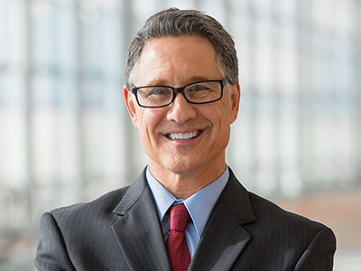 A middle-aged man wearing glasses, a dark suit, light blue shirt, and red tie, standing indoors with a blurred window background, smiling at the camera.