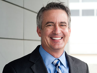 A man in a suit and tie smiles at the camera, standing in front of a modern building with horizontal lines and windows.