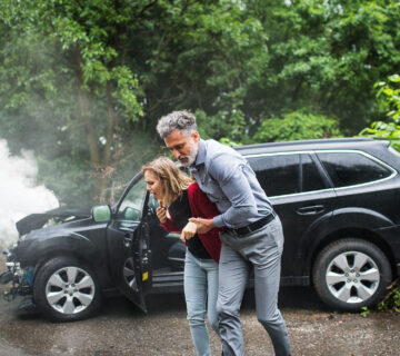 A man helps a woman away from a black SUV after an SUV rollover accident, the vehicle's front damaged and smoke rising from the hood on a roadside, surrounded by trees.