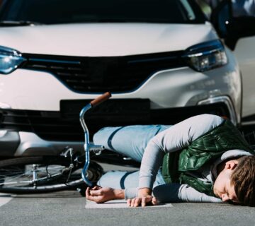 A person lies on the ground next to a fallen bicycle in front of a white car, suggesting a recent collision between the cyclist and the vehicle.