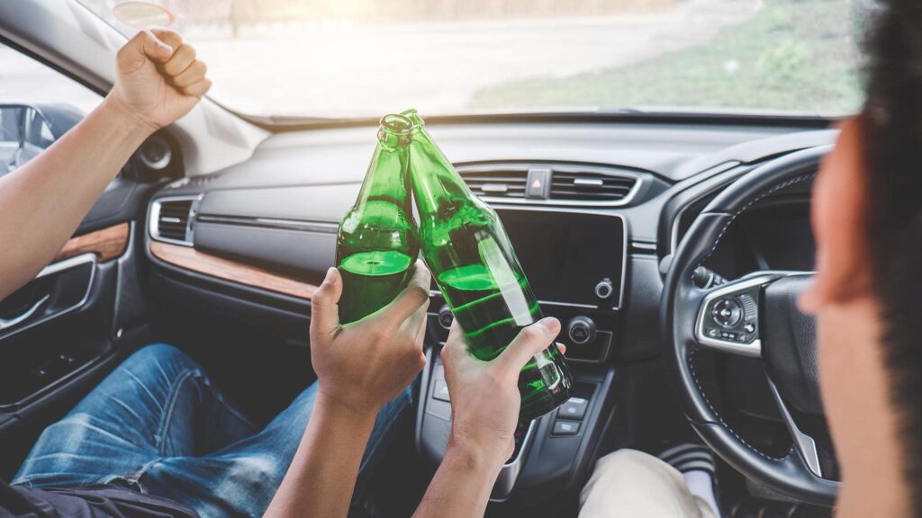 Two people sitting in a car clink green glass bottles together, with one person raising their thumb while seated in the driver's seat—an image that serves as a warning about the dangers of drunk driving accidents. - Ghassemi Law Group drunk driving accident