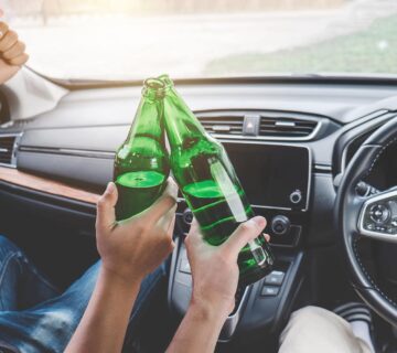 Two people sitting in a car clink green glass bottles together, with one person raising their thumb while seated in the driver's seat—an image that serves as a warning about the dangers of drunk driving accidents.