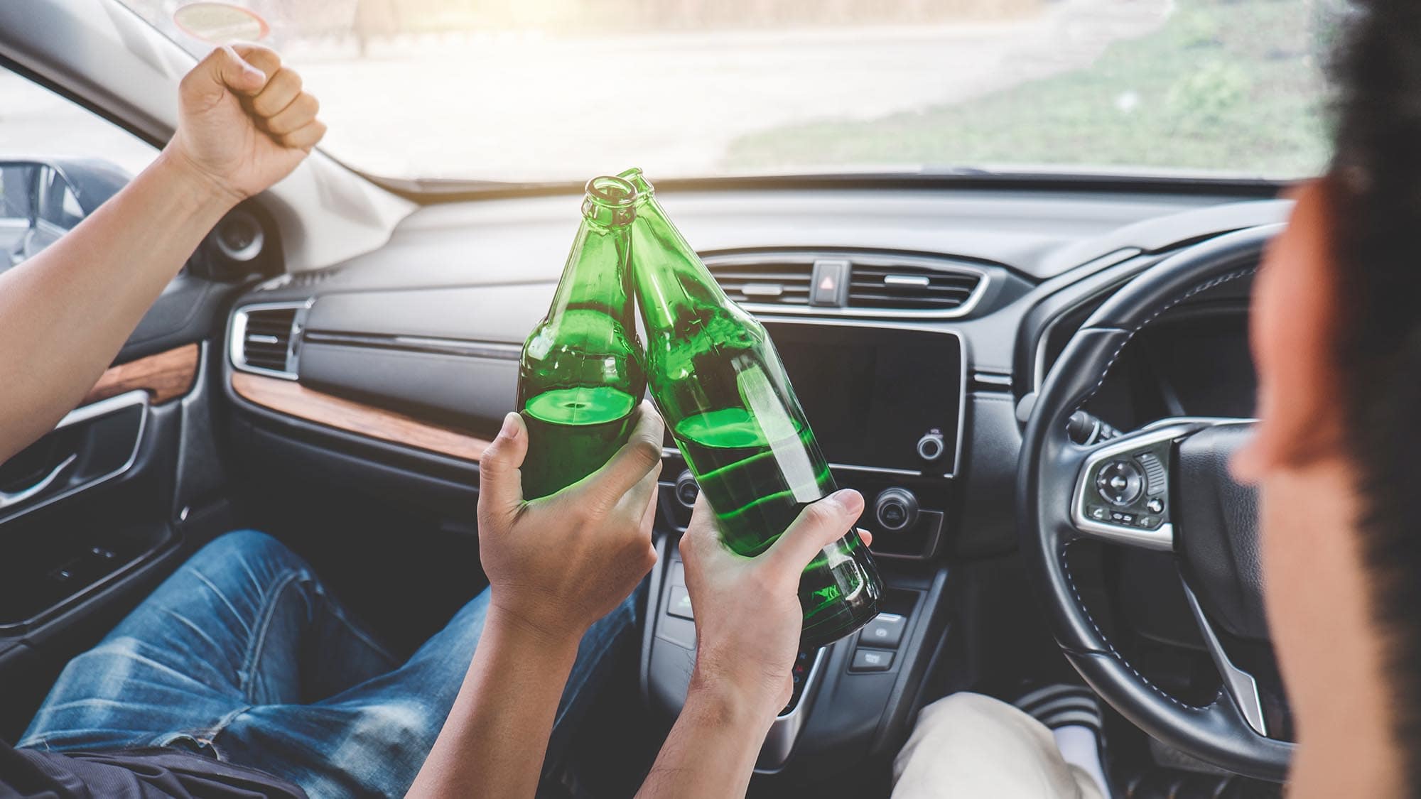 Two people sitting in a car clink green glass bottles together, with one person raising their thumb while seated in the driver's seat—an image that serves as a warning about the dangers of drunk driving accidents.