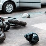 A damaged motorcycle and helmet lie on the road next to a car with an open door and broken glass, indicating a recent motorcycle accident