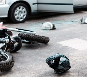 A damaged motorcycle and helmet lie on the road next to a car with an open door and broken glass, indicating a recent motorcycle accident.