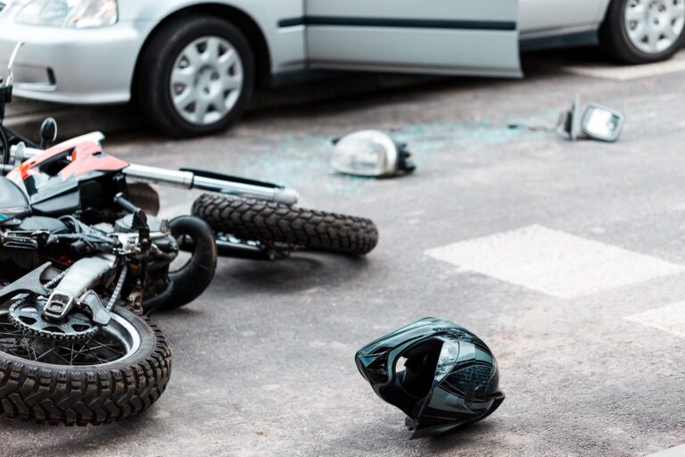 A damaged motorcycle and helmet lie on the road next to a car with an open door and broken glass, indicating a recent motorcycle accident. - Ghassemi Law Group - Ghassemi Law Group A damaged motorcycle and helmet lie on the road next to a car with an open door and broken glass, indicating a recent motorcycle accident