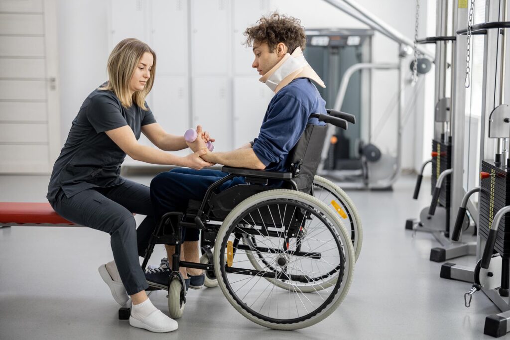 A woman assists a man in a wheelchair, who is wearing a neck brace, with a hand exercise using a small ball in a physical therapy setting.