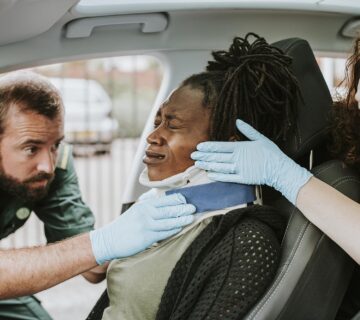 Two paramedics fit a woman with a neck brace while she sits in a car, suggesting she may have suffered a personal injury.