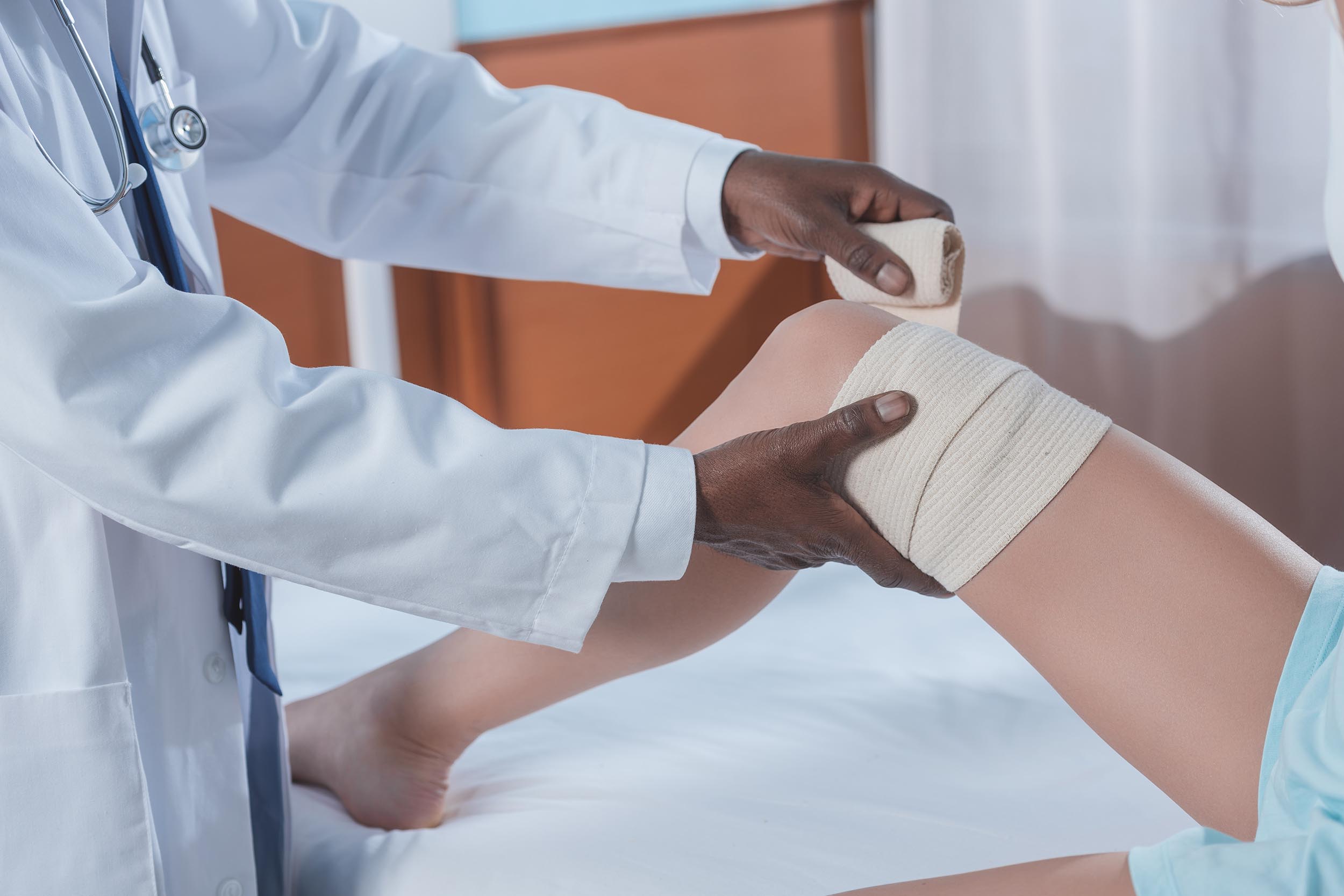 A doctor wraps a bandage around a patient's injured knee—often seen in cases of soft tissue injuries—while the patient sits on an examination table. - Ghassemi Law Group A doctor wraps a bandage around a patient's injured knee—often seen in cases of soft tissue injuries—while the patient sits on an examination table.
