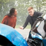 Two people stand next to damaged cars after a collision on the road, appearing to discuss the accident  Trees are visible in the background 