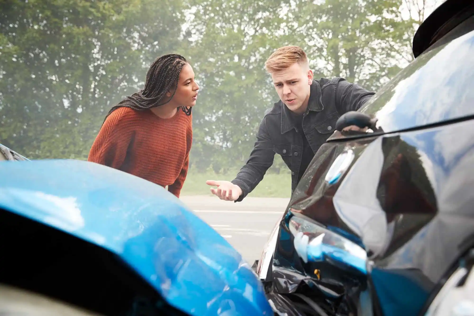 Two people stand next to damaged cars after a collision on the road, appearing to discuss the accident. Trees are visible in the background. - Ghassemi Law Group Two people stand next to damaged cars after a collision on the road, appearing to discuss the accident Trees are visible in the background