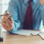 A person in business attire sits at a desk with documents, holding the scales of a Lady Justice statue