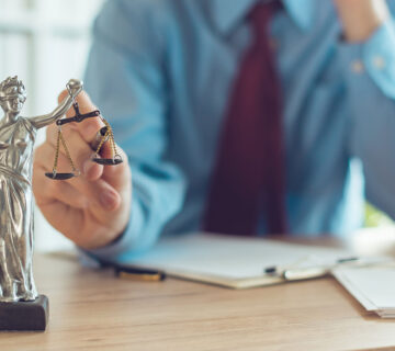 A person in business attire sits at a desk with documents, holding the scales of a Lady Justice statue.