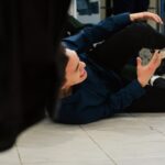 A person wearing a navy shirt and white sneakers is lying on a tiled floor, appearing to have fallen, with one arm extended and an expression of discomfort