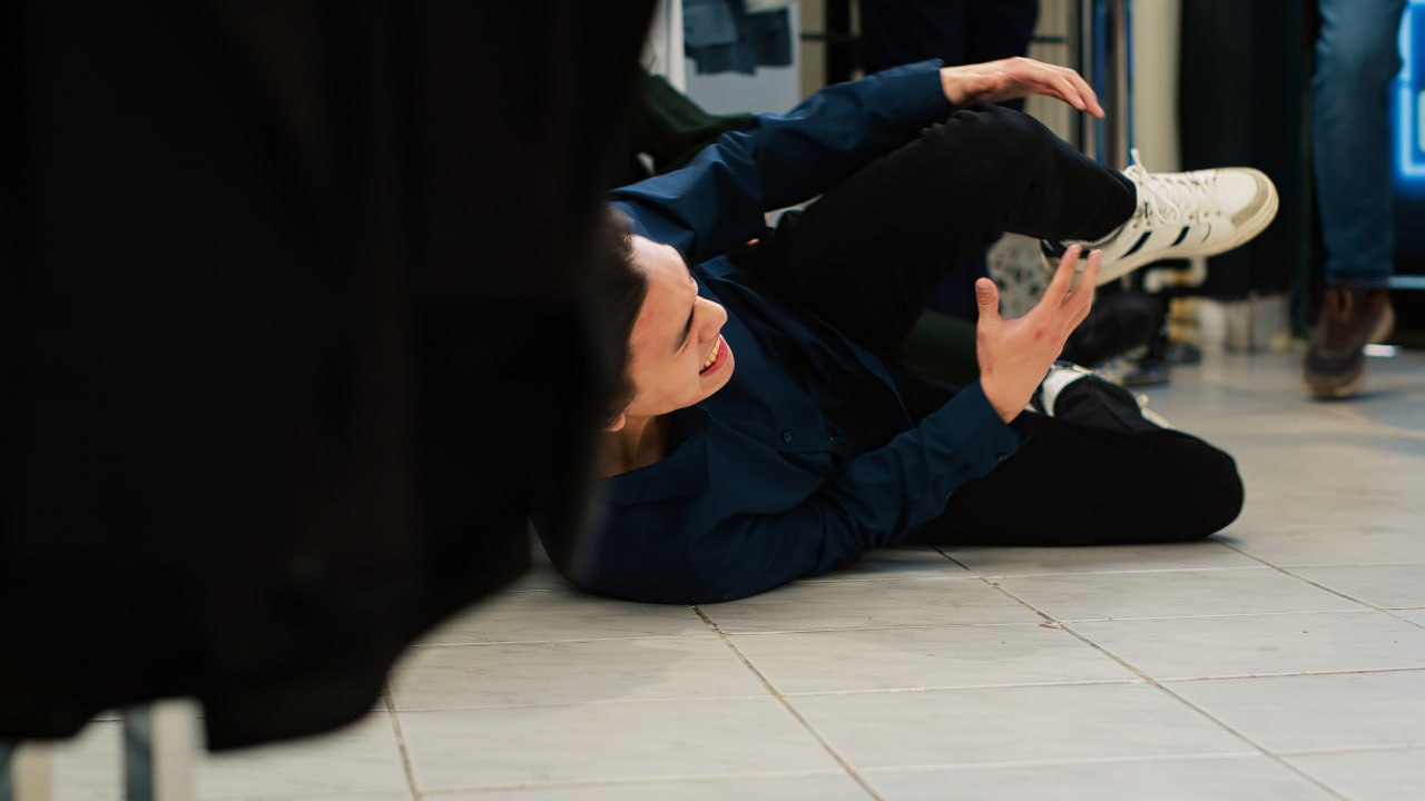 A person wearing a navy shirt and white sneakers is lying on a tiled floor, appearing to have fallen, with one arm extended and an expression of discomfort.