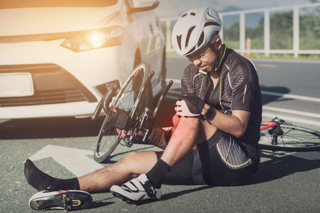 A cyclist wearing a helmet sits on the road holding his injured knee next to a fallen e-bike after an E-Bike Accident, with a white car nearby.