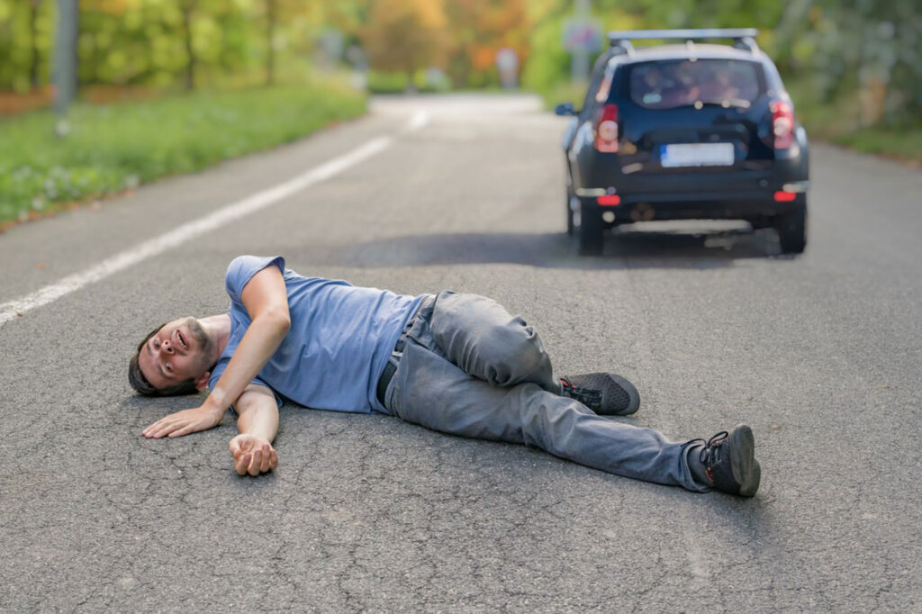 A man is lying on the road in apparent distress with a car driving away in the background on a tree-lined street. - Ghassemi Law Group A man is lying on the road in apparent distress with a car driving away in the background on a tree-lined street.