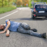 A man is lying on the road in apparent distress with a car driving away in the background on a tree lined street