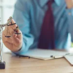 Statue of Lady Justice holding scales on a desk, with a blurred person in a suit in the background 