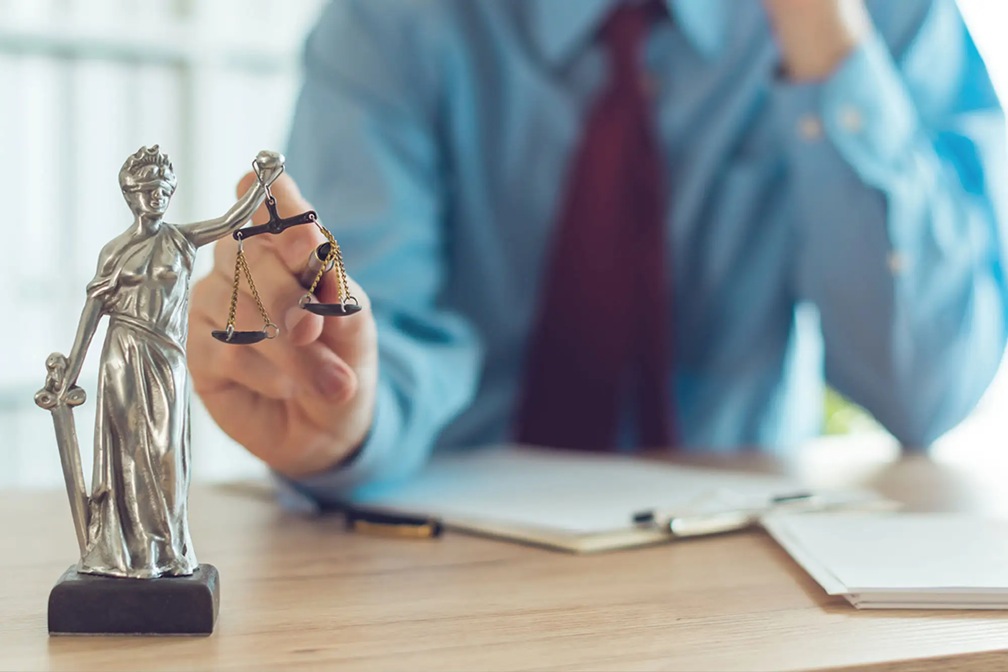 Statue of Lady Justice holding scales on a desk, with a blurred person in a suit in the background 