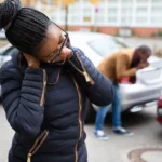 Woman holding her neck in pain after a car accident; a man inspects damage to the rear of one of the vehicles in the background 