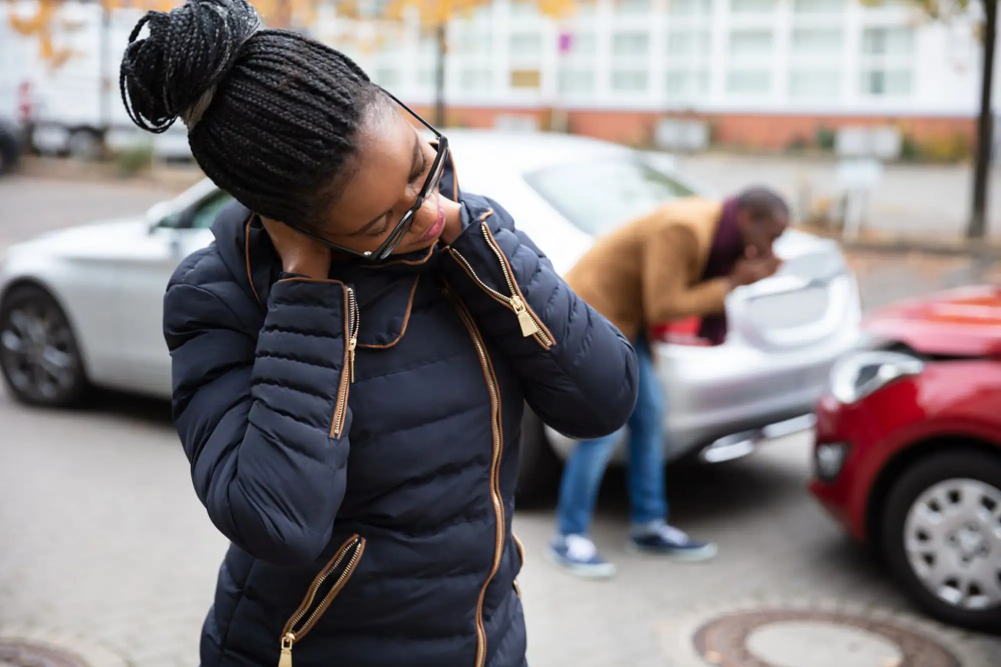 Woman holding her neck in pain after a car accident; a man inspects damage to the rear of one of the vehicles in the background 