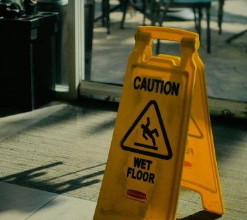 A yellow "Caution Wet Floor" sign is placed on a tiled indoor floor near a glass door, warning of possible slip and fall hazards with outdoor seating visible in the background.
