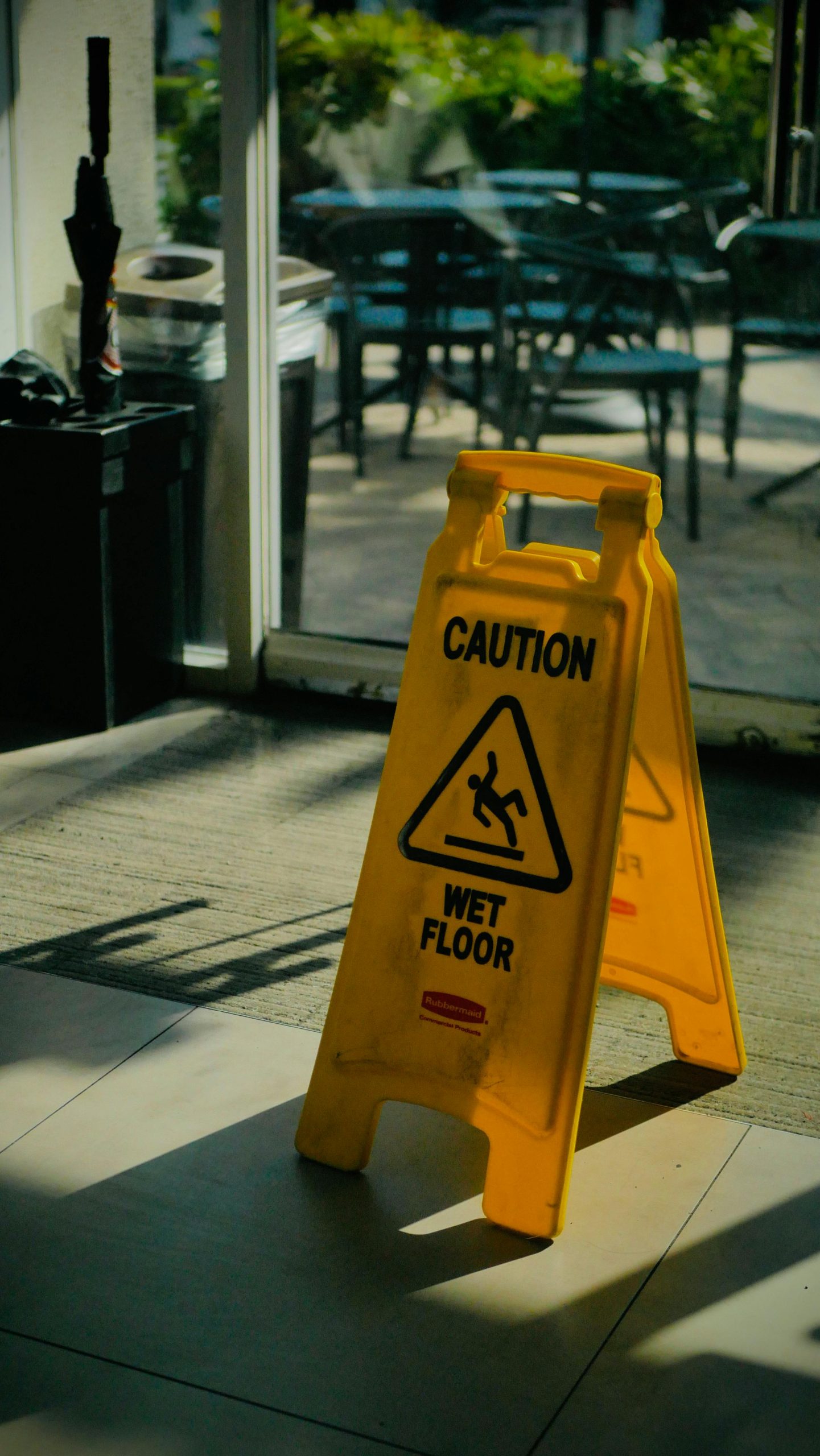 A yellow "Caution Wet Floor" sign is placed on a tiled indoor floor near a glass door, warning of possible slip and fall hazards with outdoor seating visible in the background.