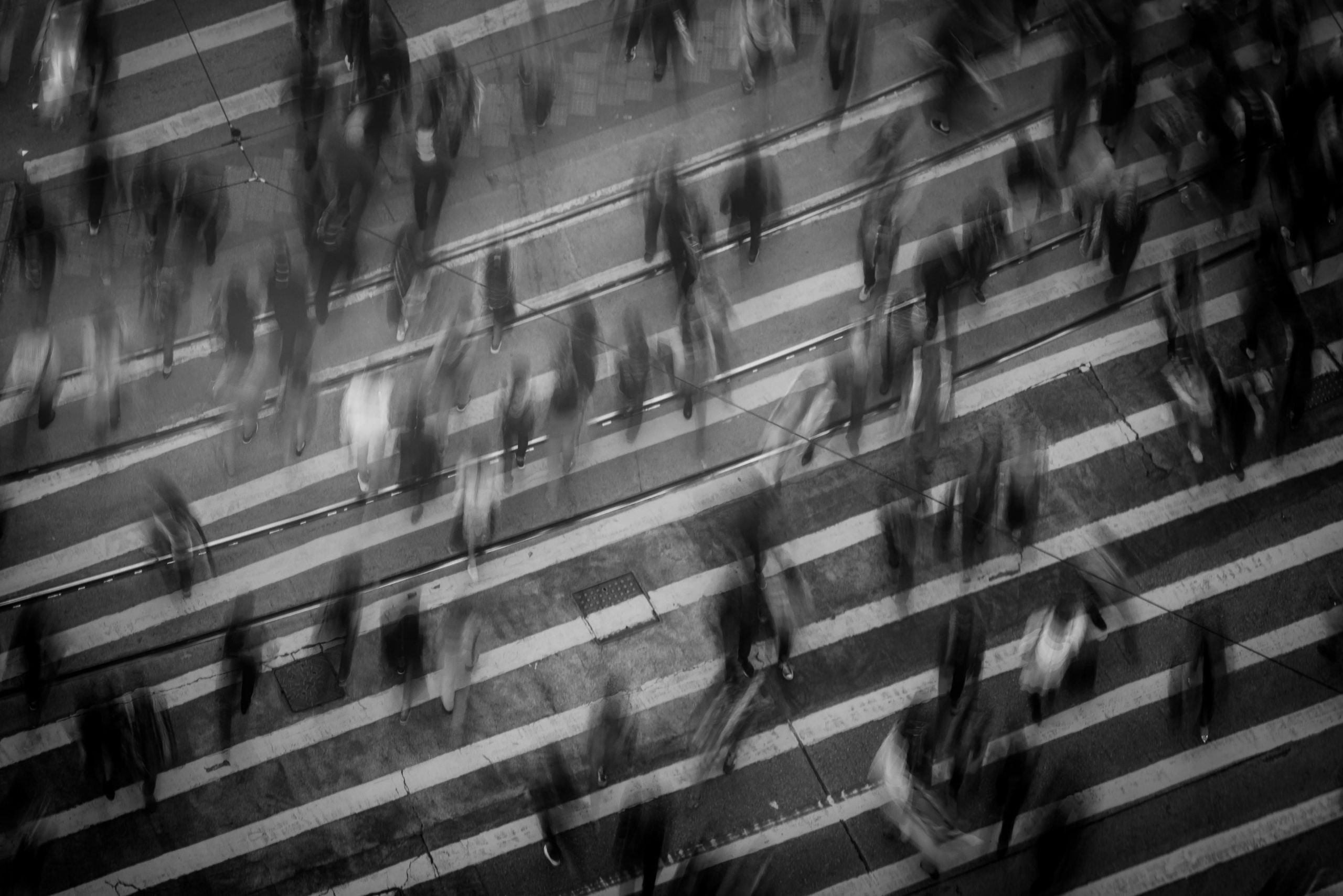 Black and white photo of a crowded crosswalk with blurred figures of people walking in different directions, evoking the chaotic potential for a pedestrian accident.