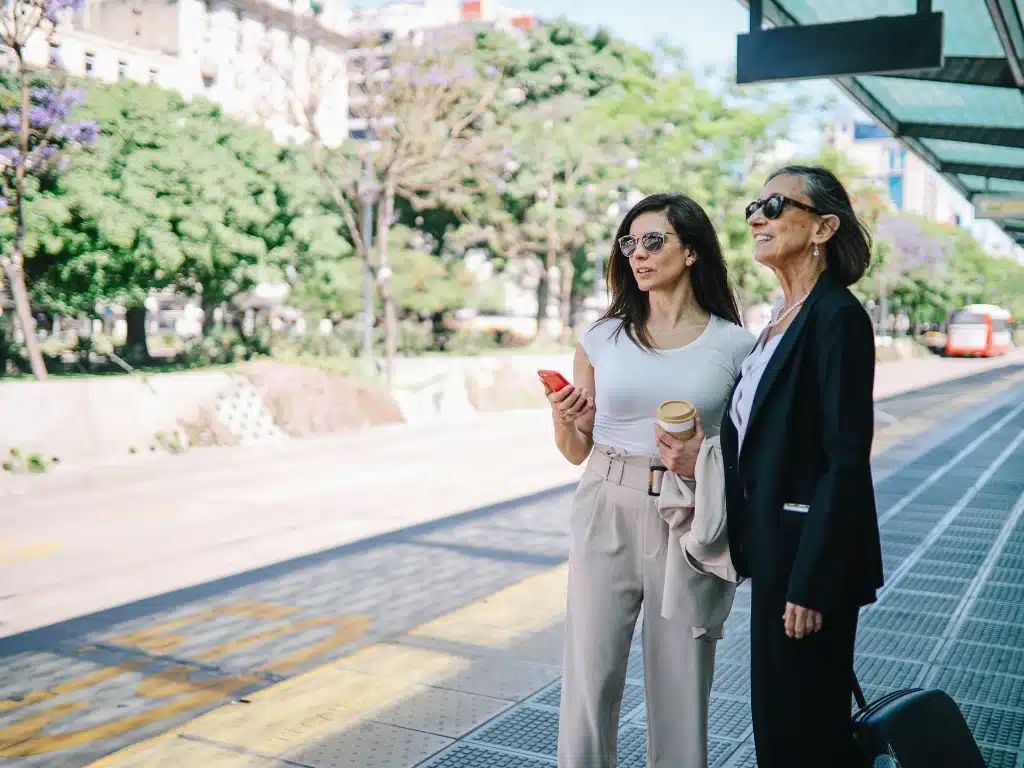 Two women in business attire stand at a city tram or bus stop, one holding a phone and coffee cup, the other with a suitcase, waiting in an urban outdoor setting. - Ghassemi Law Group Two women in business attire stand at a city tram or bus stop, one holding a phone and coffee cup, the other with a suitcase, waiting in an urban outdoor setting.
