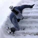 Person slipping and falling on snow covered outdoor steps, wearing winter clothes and boots, surrounded by snow 