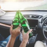Two people in a car clink green glass bottles in a toast, with the dashboard and steering wheel visible 