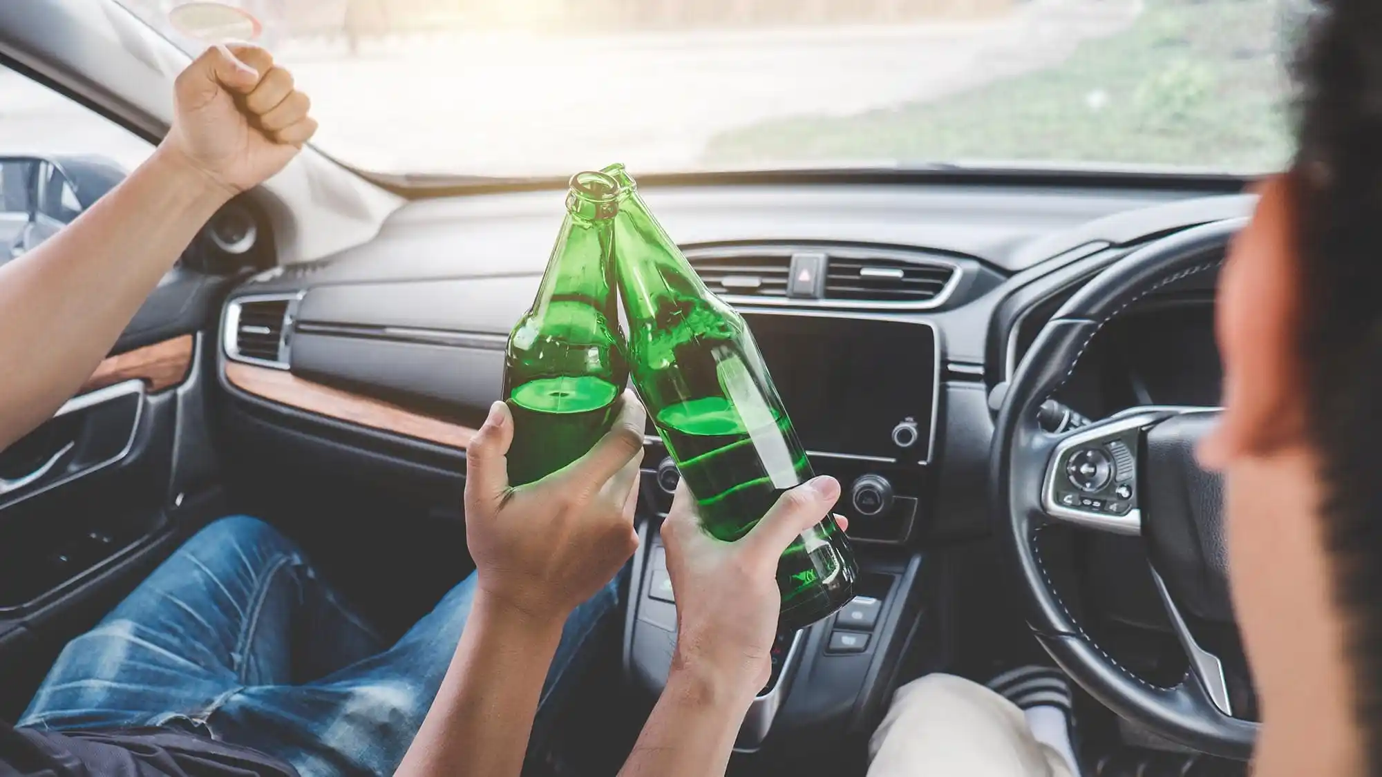 Two people in a car clink green glass bottles in a toast, with the dashboard and steering wheel visible.
