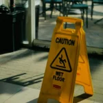 A yellow caution sign reading “Caution Wet Floor” stands indoors, warning of a slippery surface Tables and chairs are visible outside through a window