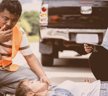 Two people in safety vests assist an unconscious person lying on the ground near a pickup truck; one uses a radio and the other takes notes.