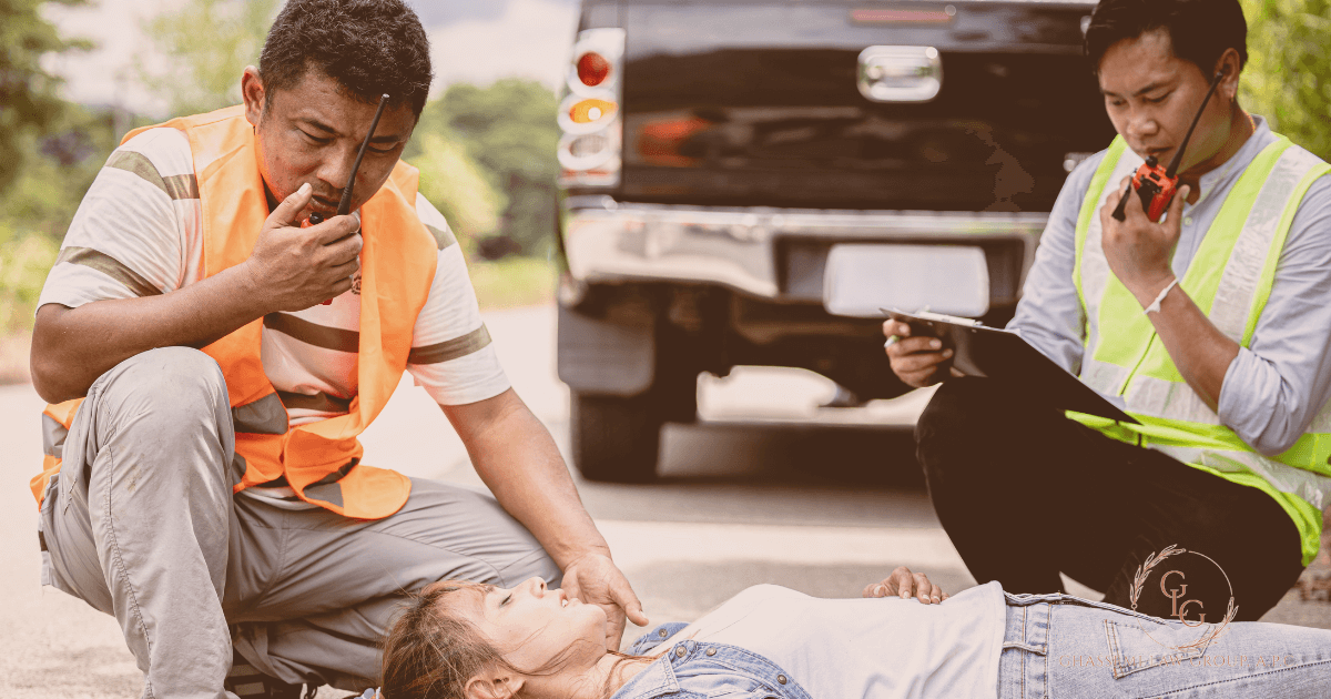 Two people in safety vests assist an unconscious person lying on the ground near a pickup truck; one uses a radio and the other takes notes.