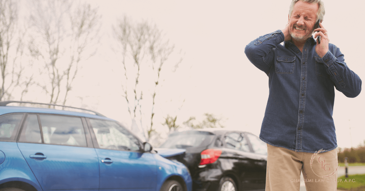 A man stands holding his neck and talking on the phone beside two damaged cars after a rear end collision.