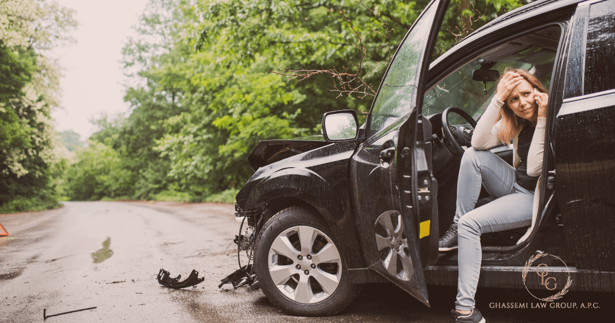 A woman sits in the open door of a black car with front end damage on a deserted road, looking distressed and holding her head.