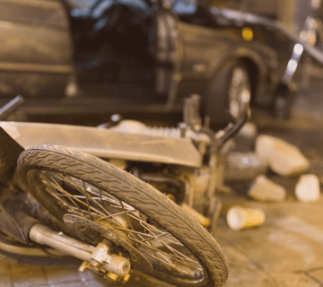 A damaged motorcycle lies on its side near scattered debris and a car with an open door, suggesting a recent accident on a city street.