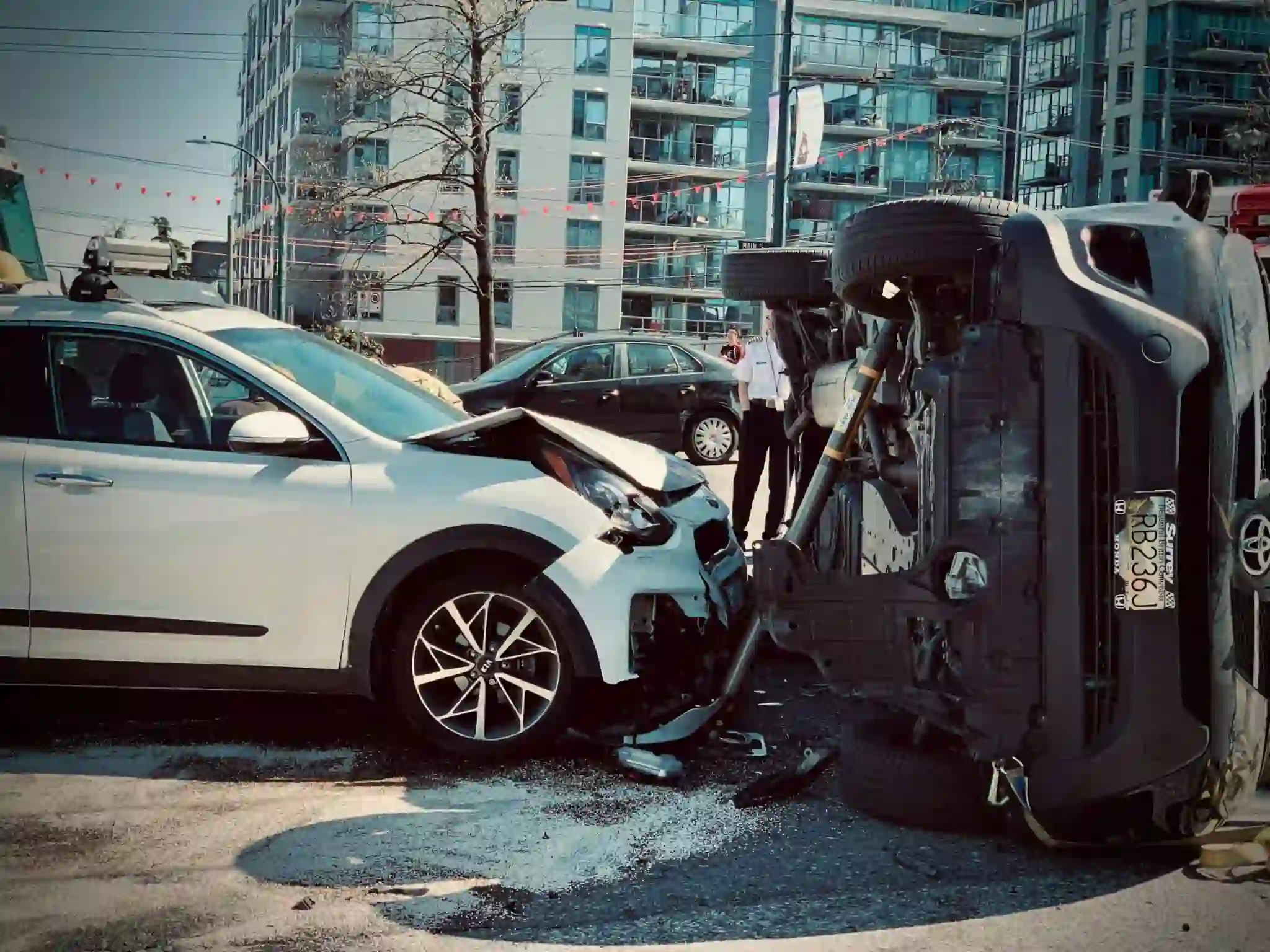 A white car and a black SUV are involved in a collision; the SUV is flipped on its side, with visible damage. An onlooker and city buildings are in the background. - Ghassemi Law Group A white car and a black SUV are involved in a collision; the SUV is flipped on its side, with visible damage An onlooker and city buildings are in the background