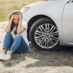 A woman sits on the ground next to a silver car with her head in her hands, appearing distressed  The car’s hood is open and one tire is visible, suggesting trouble has found her even in scenic Laguna Beach 