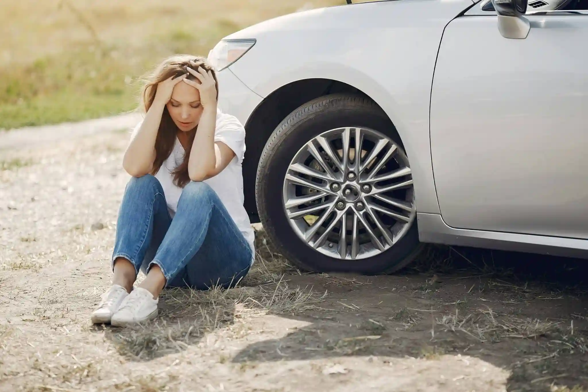 A woman sits on the ground next to a silver car with her head in her hands, appearing distressed  The car’s hood is open and one tire is visible, suggesting trouble has found her even in scenic Laguna Beach 
